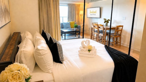 Ein Hotelzimmer mit einem Bett, einem Nachttisch mit Blumenvase und Blick auf den Wohnbereich mit einer Couch und einem Fernseher, der 'Instant Suites' anzeigt.
