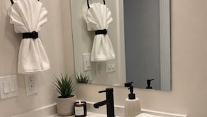 Ein modernes Badezimmer mit einem weißen Waschbecken, einem schwarzen Wasserhahn und einem Spiegel darüber. Zwei Handtücher hängen an schwarzen Handtuchringen, ein kleiner Topfpflanze und ein Toilettenpapierhalter sind ebenfalls vorhanden.