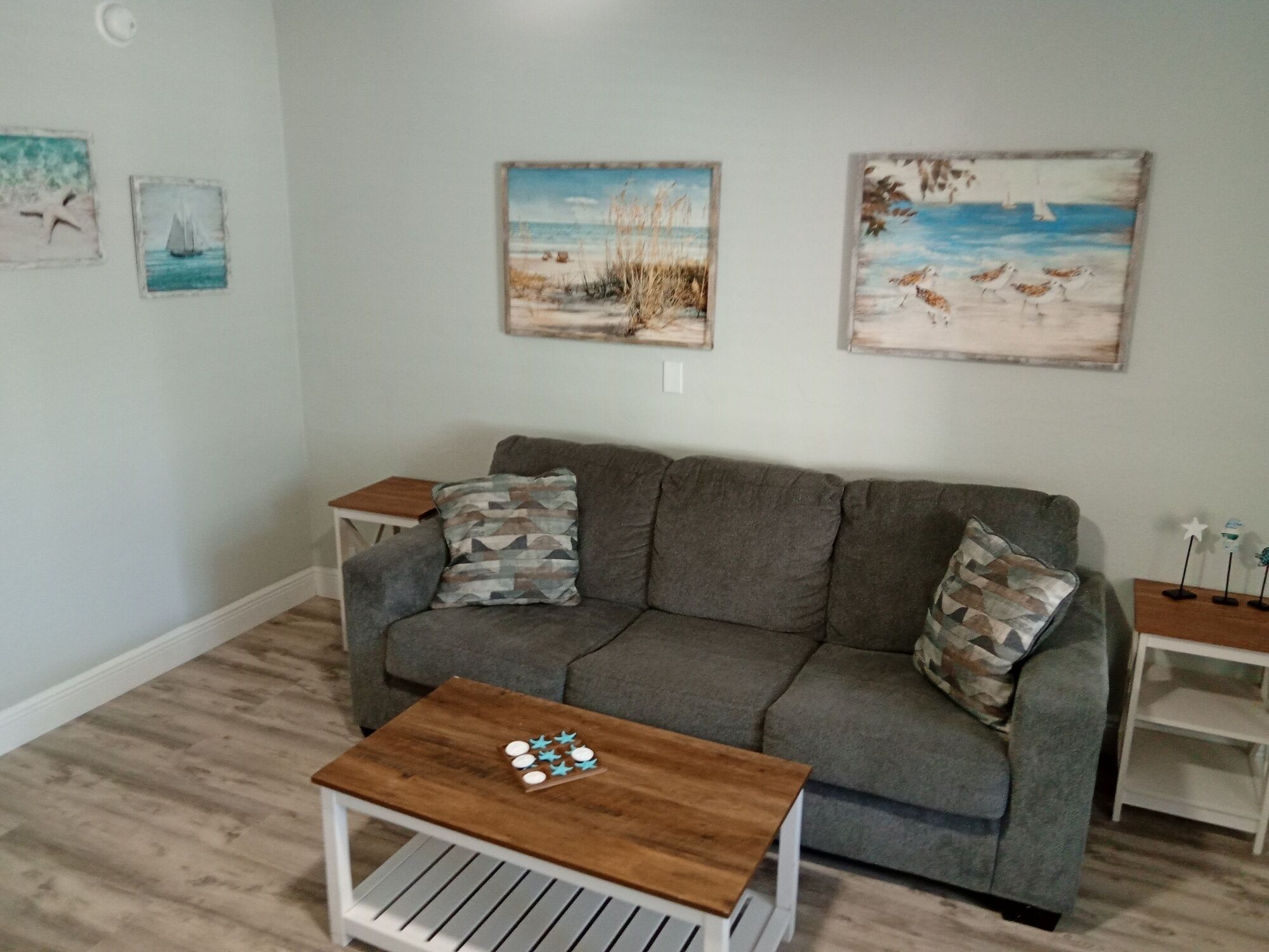 A living room with a gray sofa, two side tables, a coffee table, and wall art depicting beach scenes.