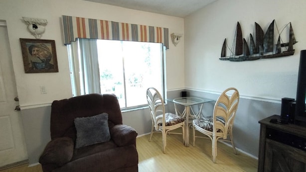 Ein Wohnzimmer mit einem braunen Sessel, einem Esstisch mit Glasplatte und zwei Stühlen, einem Fernseher und einer Wanddekoration.