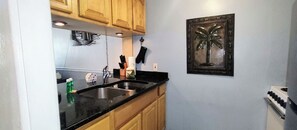 A kitchen with wooden cabinets, a black countertop, a sink, a stove, and a wall-mounted picture of a palm tree. - Unit 113 - The Captain's Bayside Getaway (2nd Floor) (Treasure Island)