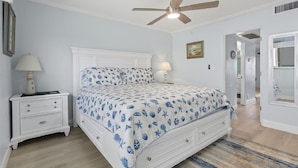 Quarto com cama, mesas de cabeceira, ventilador de teto e porta para outro cômodo.