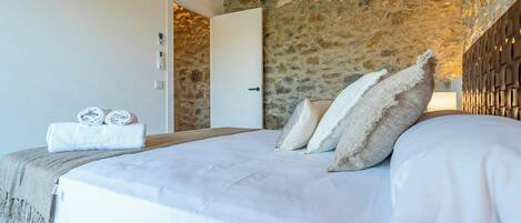 A bedroom with a stone wall, a bed with white bedding, and a door leading to another room.
