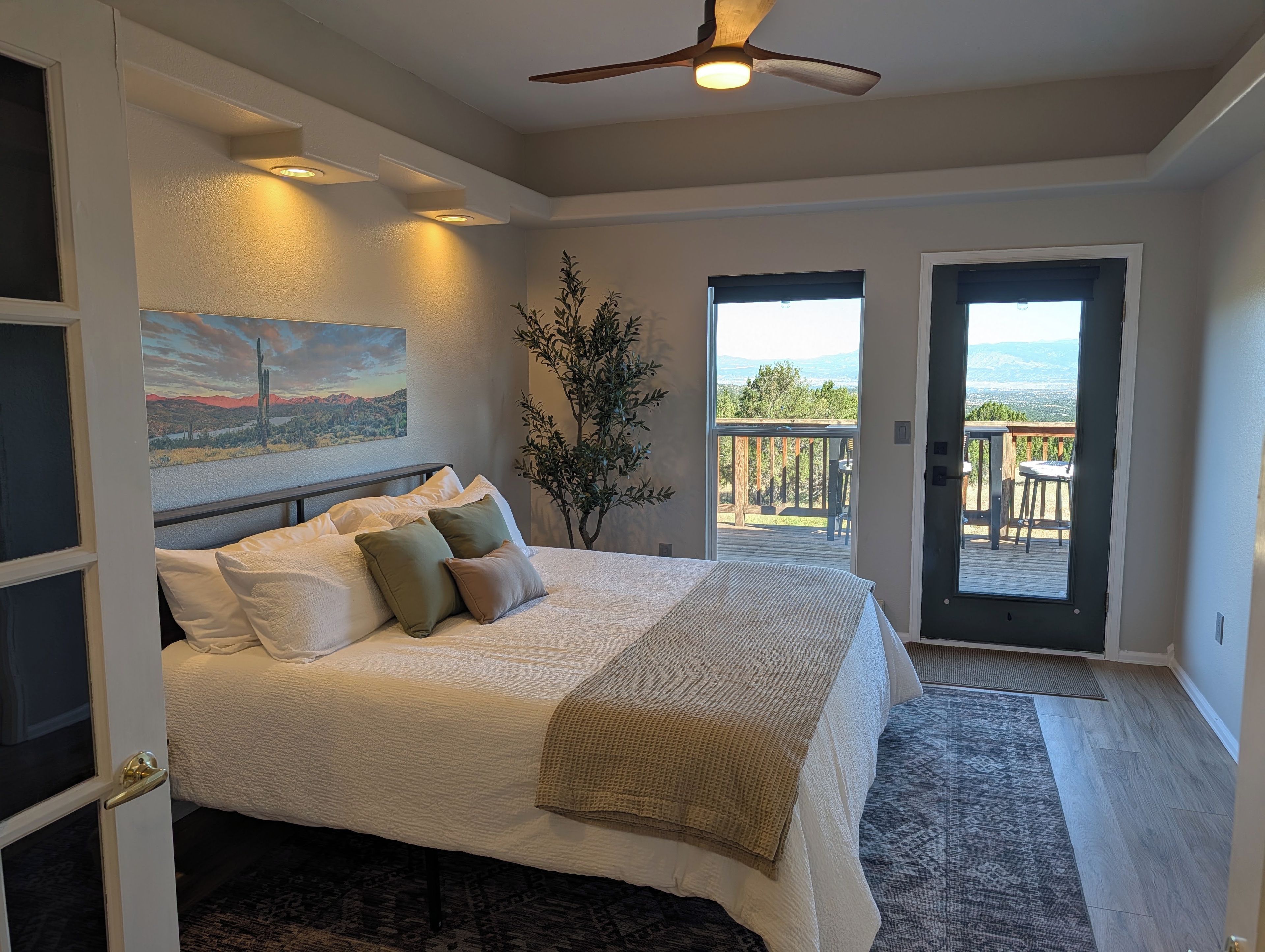Une chambre à coucher avec un lit, un ventilateur de plafond, un tableau, une plante et une vue sur l’extérieur, visible à travers une porte.