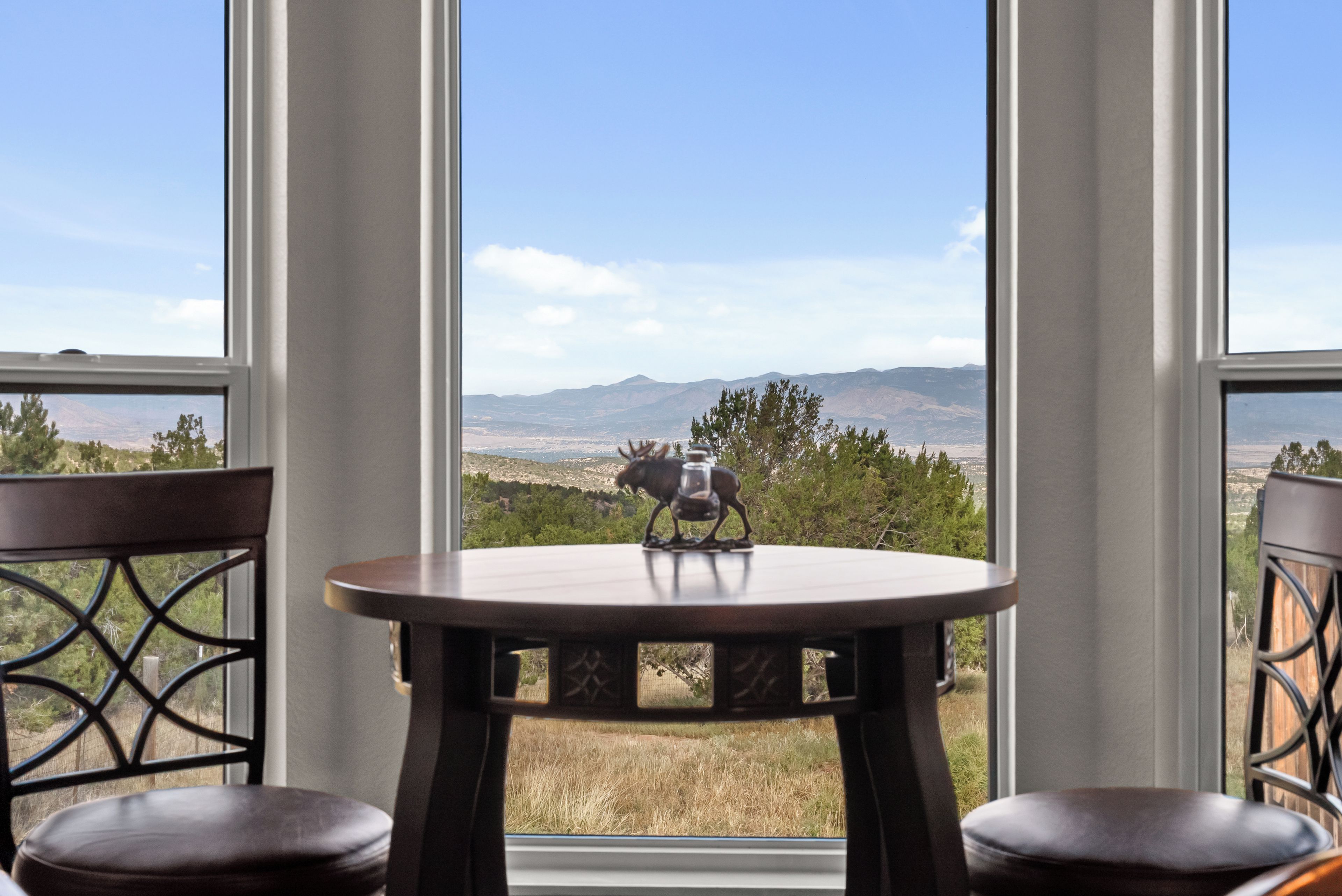 A table with a decorative item in the center, two chairs on either side, and a view of a mountainous landscape through large windows.