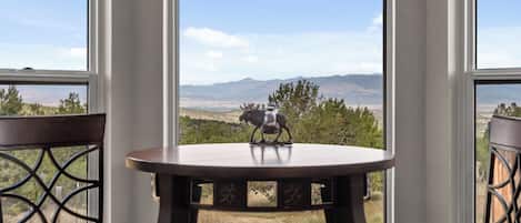 A table with a decorative item in the center, two chairs on either side, and a view of a mountainous landscape through large windows.