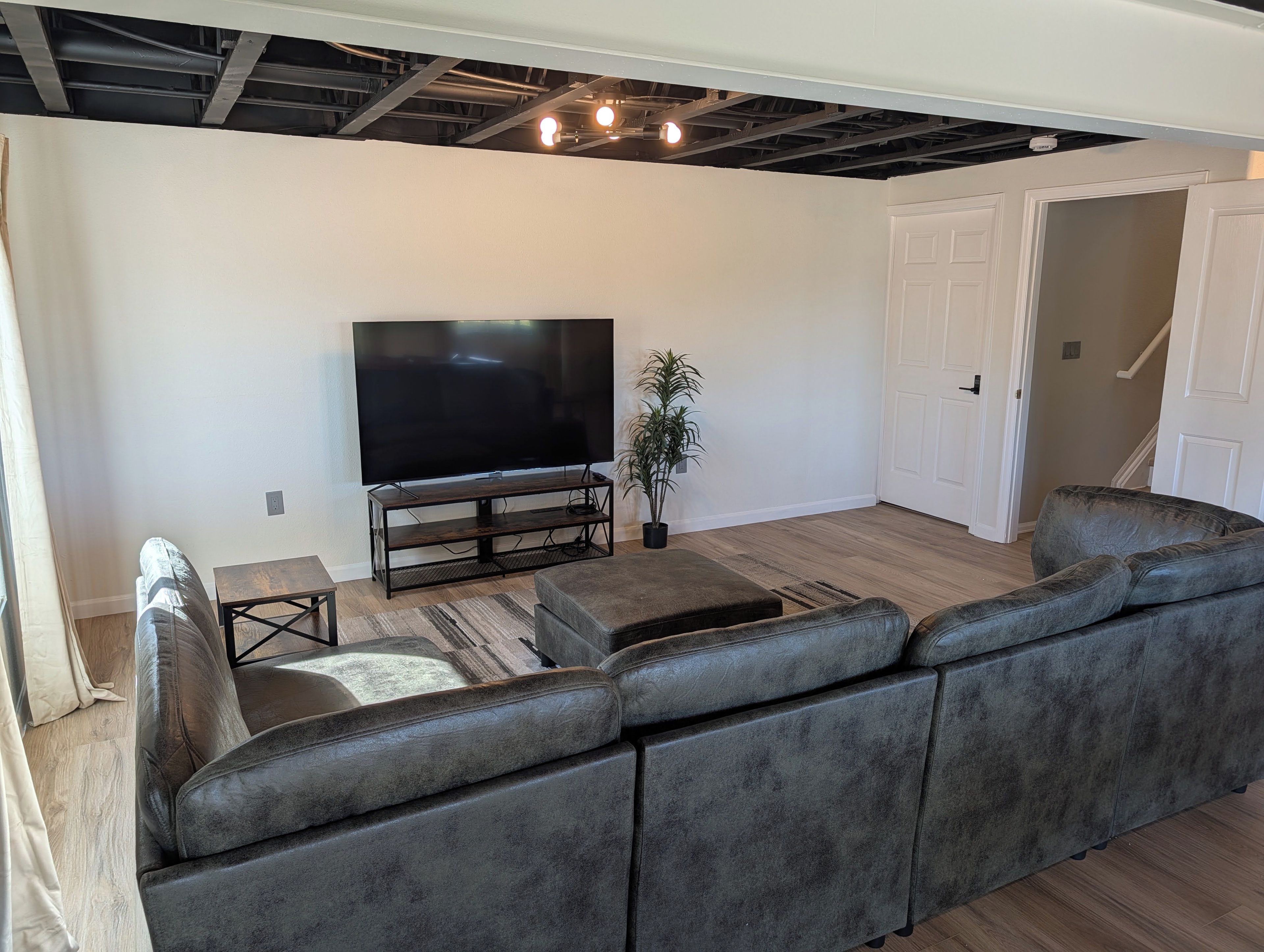 A modern living room with a large flat-screen TV, a dark wood coffee table, a grey sectional sofa, and a potted plant.