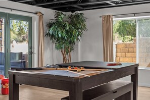 A billiards table in a room with a large plant, a window with curtains, and a door.