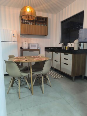 A modern kitchen with a dining table, chairs, a hanging light fixture, a microwave, and a refrigerator. - Cozy apartment on Coroa Vermelha Beach (Porto Seguro)