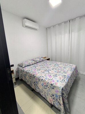 A bedroom with a bed, a nightstand, a wall-mounted air conditioner, and a window with curtains. - Cozy apartment on Coroa Vermelha Beach (Porto Seguro)