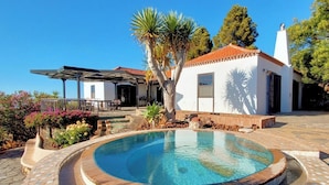 Zona de piscina amb una piscina rodona, un edifici blanc amb teulada vermella i un porxo cobert amb mobles d'exterior.