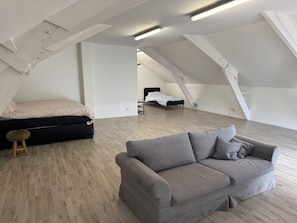 Amplo quarto no estilo loft com um sofá cinzento grande, cama com cobertura branca e piso de madeira.