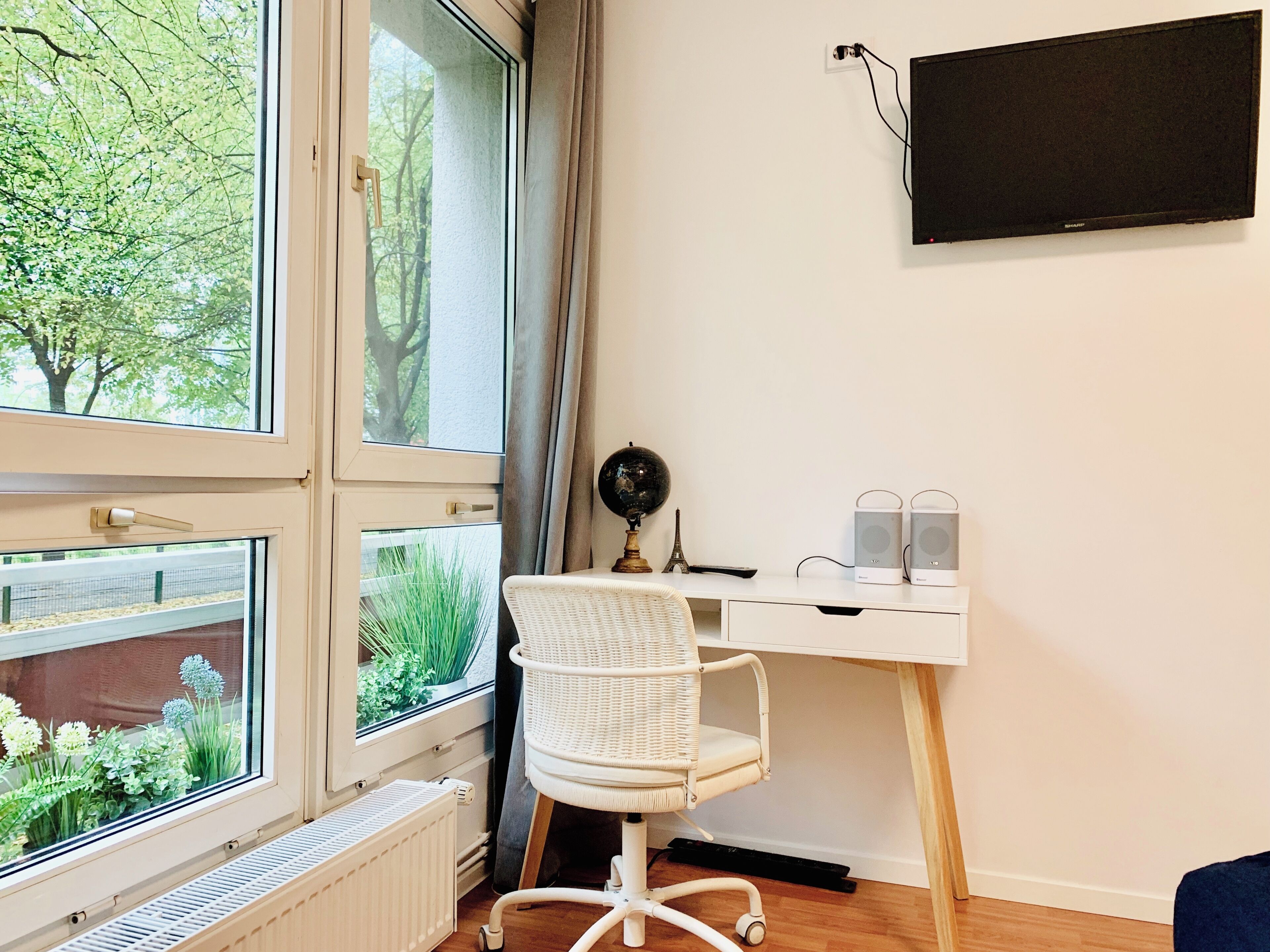 Ein Zimmer mit Schreibtisch, Stuhl, Heizkörper und einem großen Fenster mit Blick auf Grünflächen.