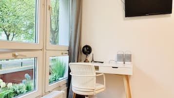Ein Zimmer mit Schreibtisch, Stuhl, Heizkörper und einem großen Fenster mit Blick auf Grünflächen.