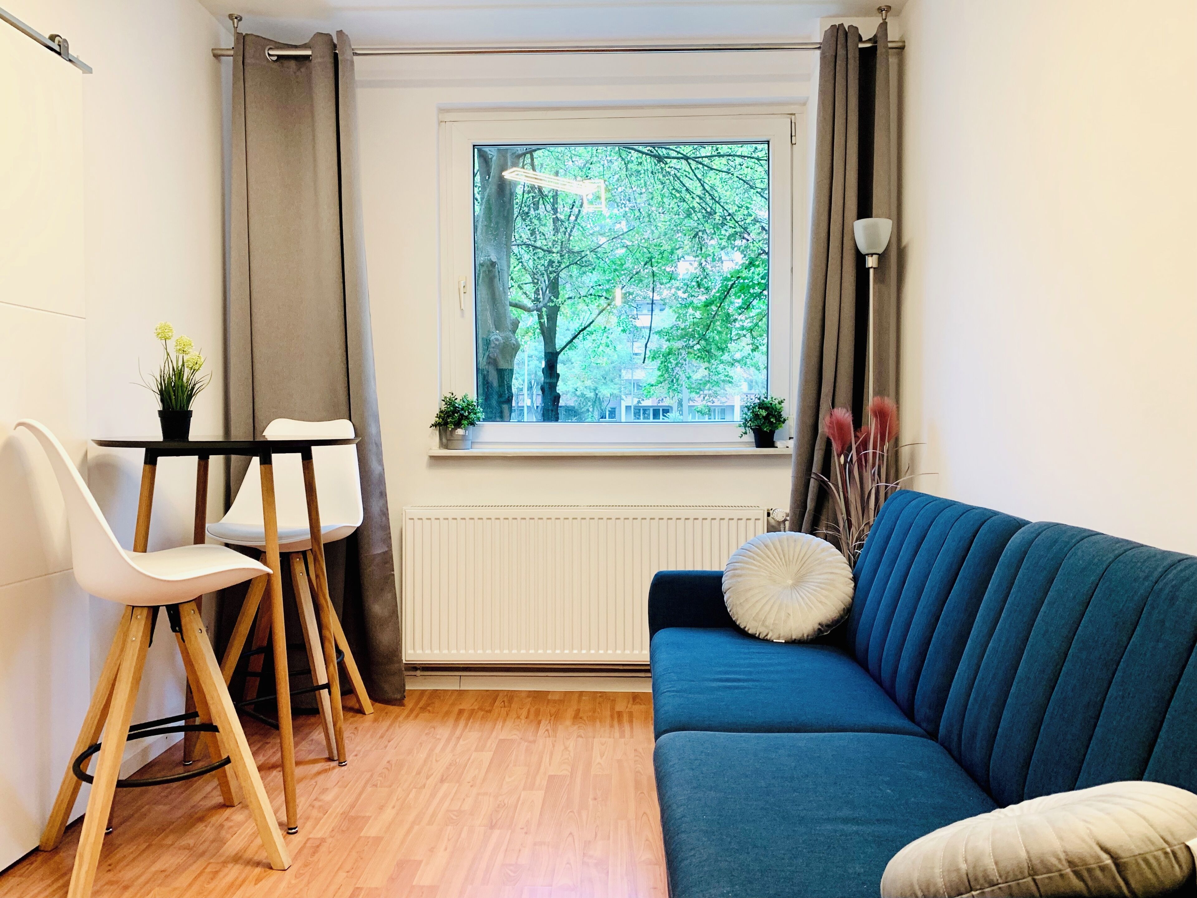 Una habitación con un sofá azul, una silla blanca, una mesita, una ventana con vistas a árboles y un radiador.