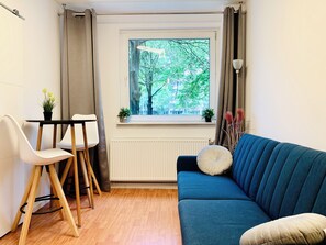 Ein Zimmer mit einem blauen Sofa, einem weißen Sessel, einem kleinen Tisch, einem Fenster mit Blick auf Bäume und einem Heizkörper.