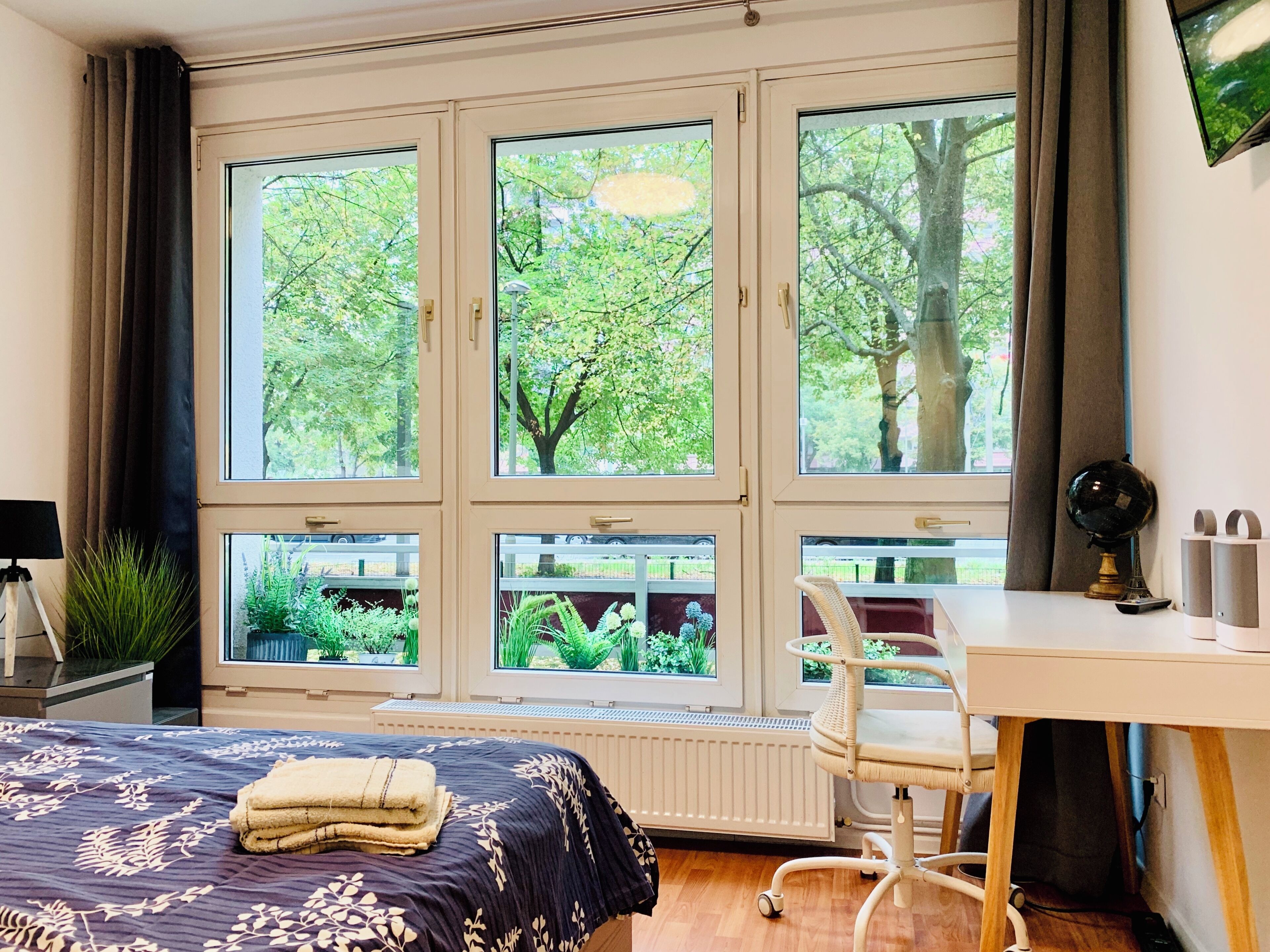 Ein Schlafzimmer mit großem Fenster, einem Bett mit Blumenmuster, einem Schreibtisch mit Stuhl und einem Heizkörper.