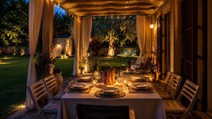 Ein Essbereich im Freien mit einem überdachten Tisch für vier Personen, umgeben von Stühlen, mit Blick auf einen Garten und ein Haus im Hintergrund.