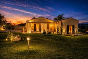 Une maison à deux étages avec une façade en pierre, un patio couvert et une pelouse bien éclairée au crépuscule.