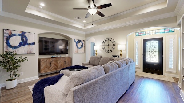 A modern living room with a sectional sofa, a flat-screen TV, decorative wall art, a large clock, and a door with a stained glass window.