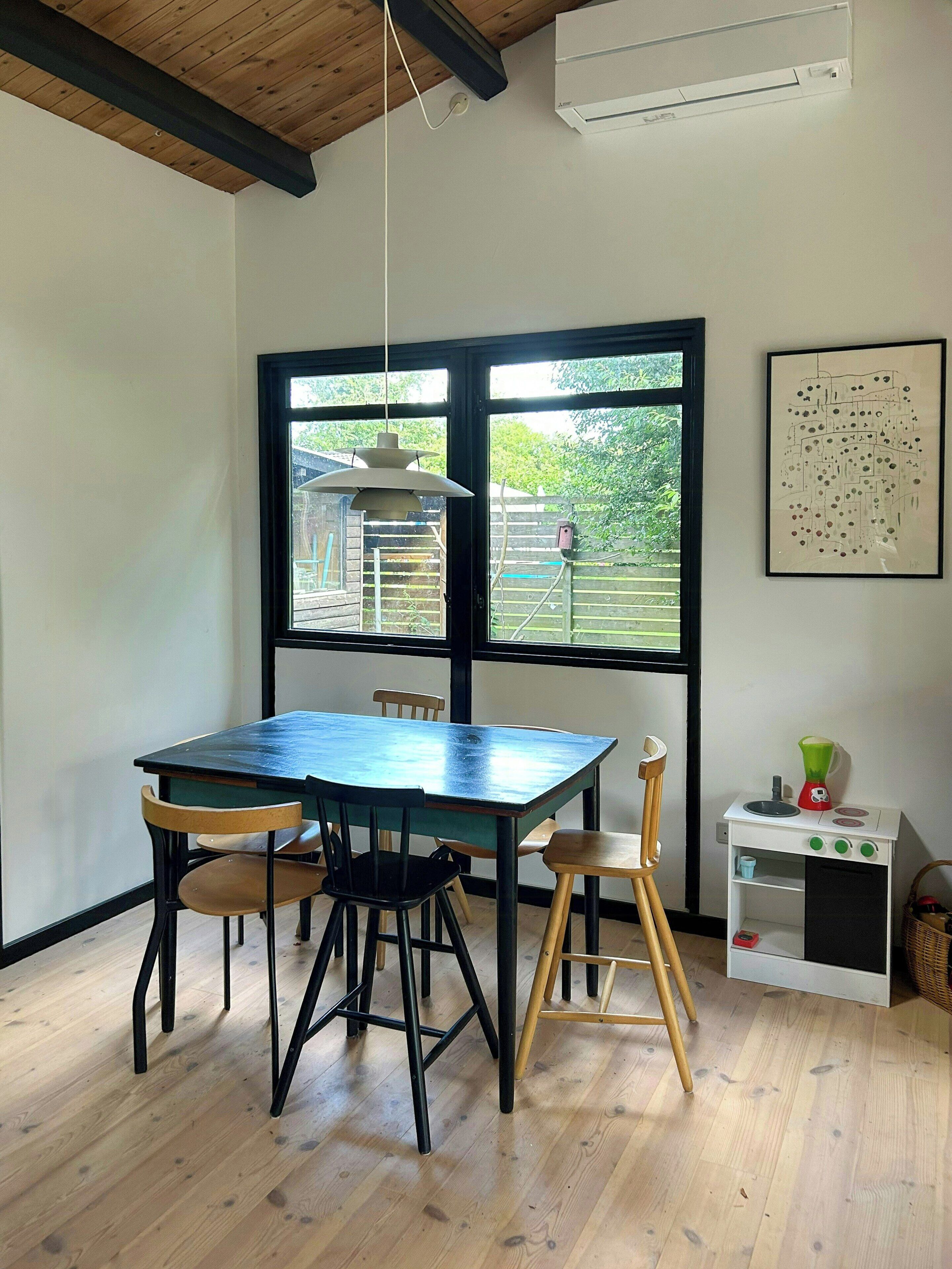 A dining area with a table and chairs, a window with a view of greenery, and a wall-mounted air conditioner.
