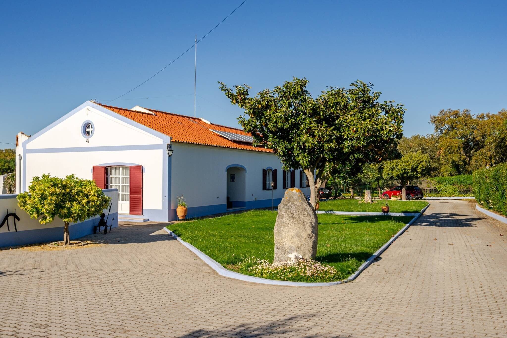 A white building with a red-tiled roof, a paved driveway, and a grassy area with a large rock.