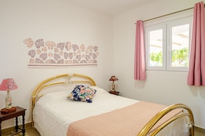 A bedroom with a bed, a nightstand, a lamp, a window with curtains, and a wall decoration. - Cottage 'Casa Dos Girassóis' with Shared Pool, Wi-Fi and Air Conditioning (Évora)