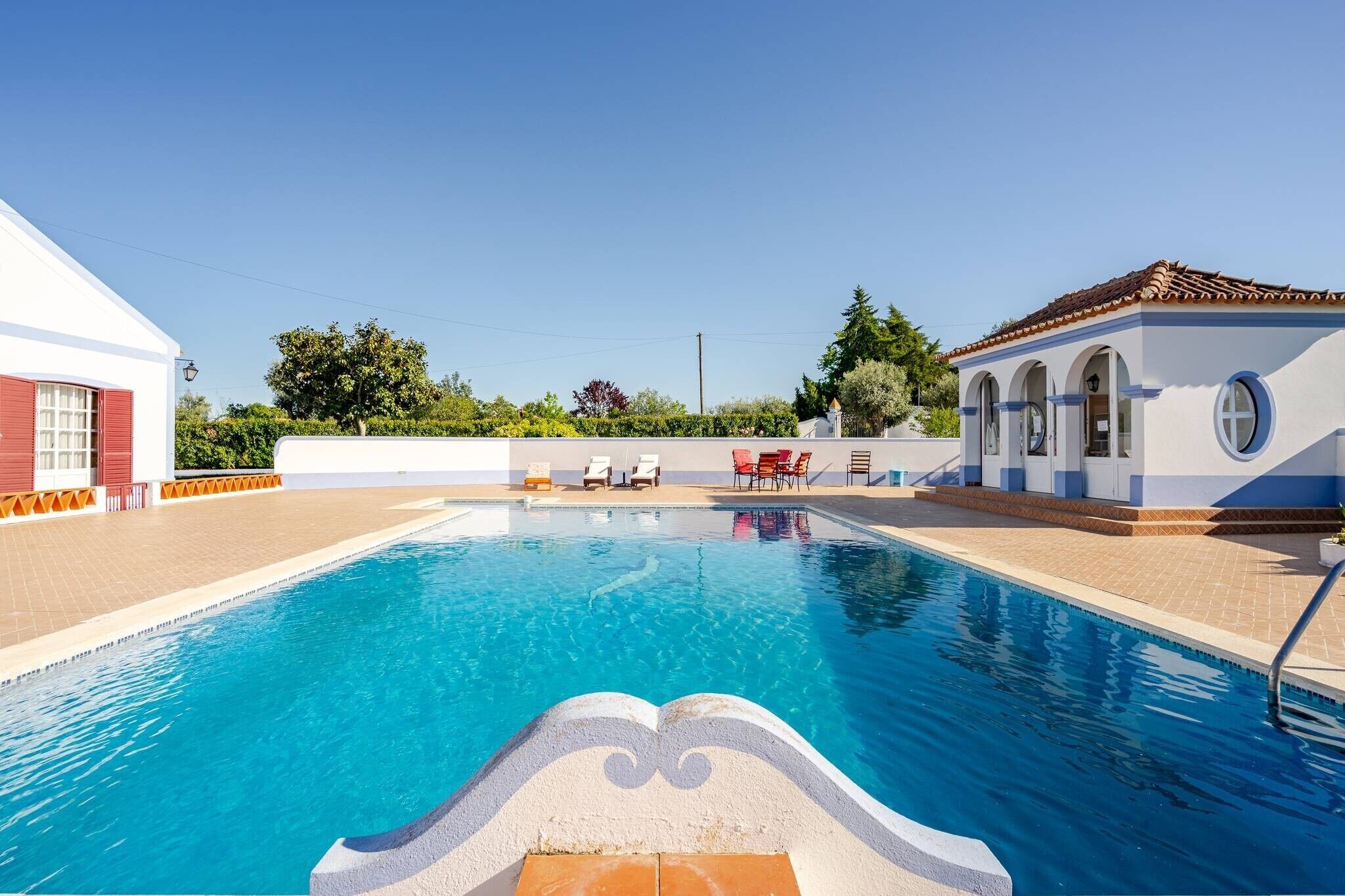 A swimming pool with clear blue water, surrounded by a white building with red shutters, and a small structure with a tiled roof.