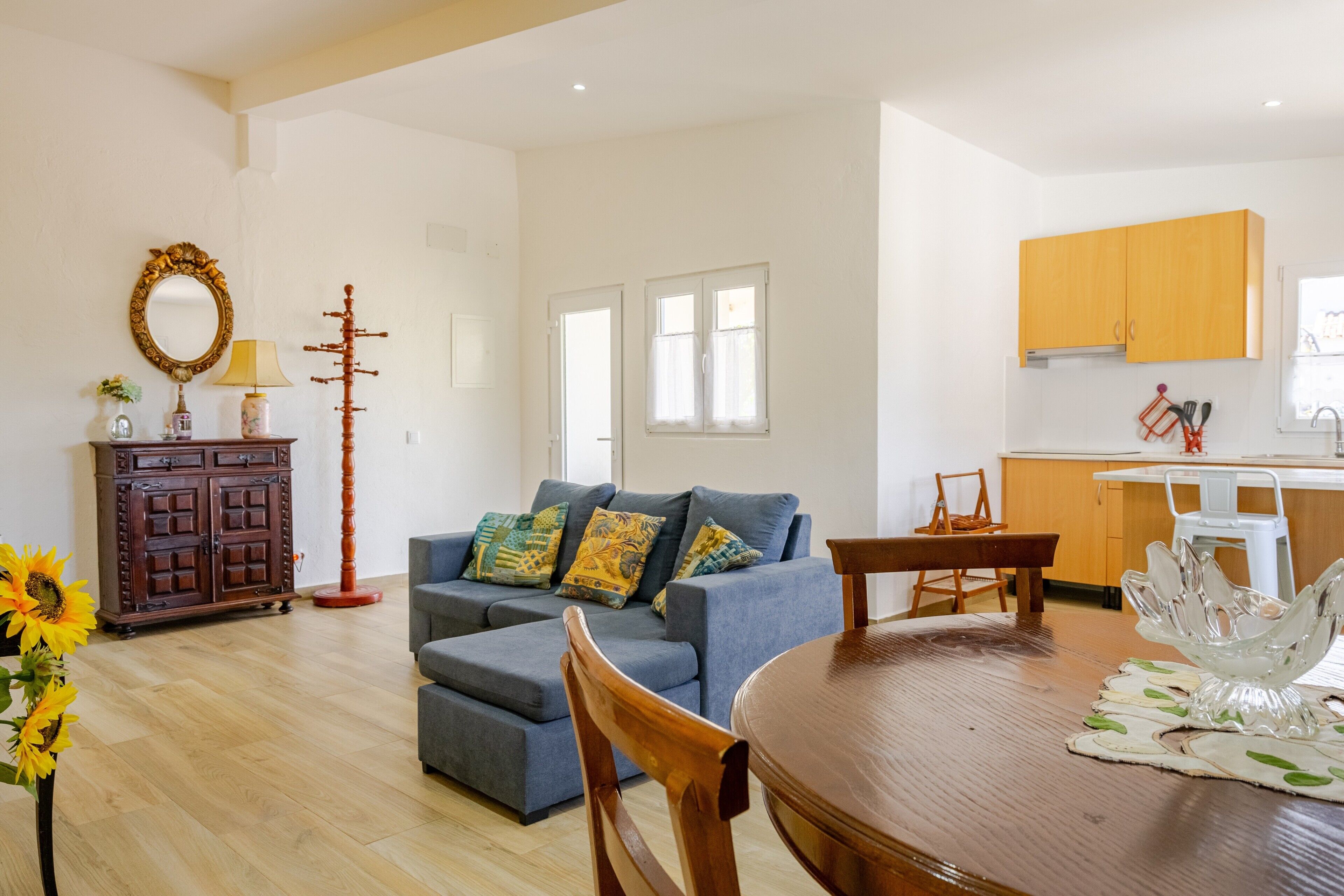 A living room with a sofa, ottoman, and a dining table with chairs. There is a kitchen area with cabinets and a window.