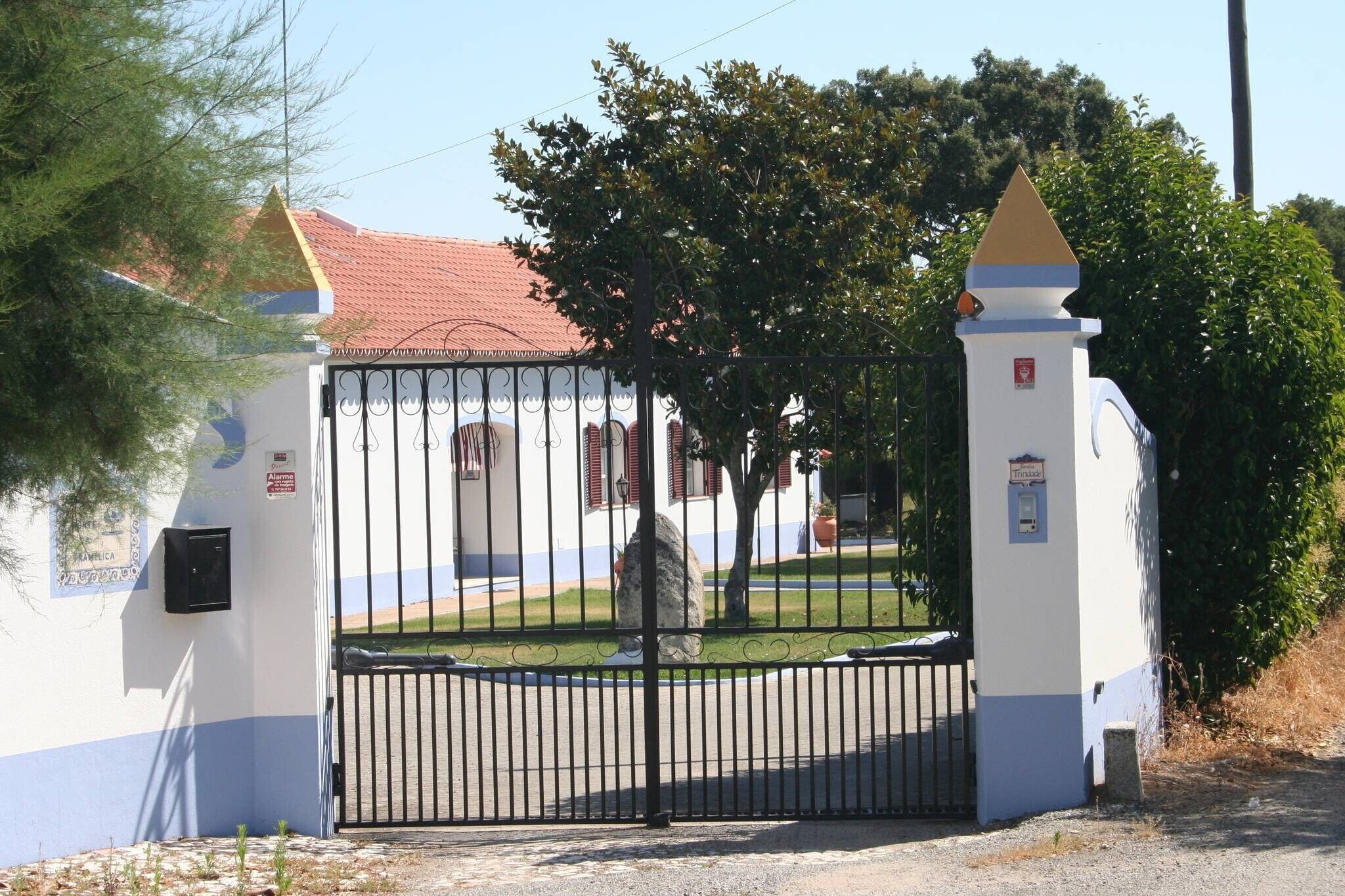 A gated entrance with a metal gate, a white building with a red roof, and a tree in the background.