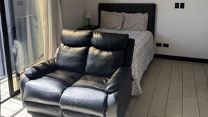 Un dormitorio moderno con un sofá de cuero negro, una cama con ropa de cama estampada y un ventanal de vidrio que da a un espacio exterior.