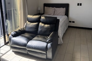 Een moderne slaapkamer met een zwarte leren bank, een bed met beddengoed met een patroon en een glazen deur naar een buitenruimte.