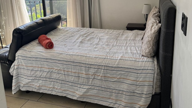 Un dormitorio con cama, un sillón de cuero negro, una mesita de noche con lámpara y una ventana con cortinas.