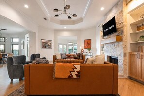 A modern living room with a sectional sofa, a fireplace, and a flat-screen TV mounted on a stone wall. - The Canyon Social - Pool - Hot Tub - Sport Court (Dripping Springs)