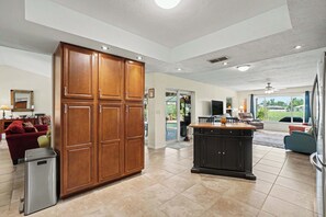 A spacious living area with a large wooden cabinet, a kitchen island, and a seating area with a red sofa and a blue chair. - Back Porch Social | Pool, Waterfront, Tiki Bar (Cape Coral)