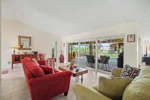 A spacious living room with a red sofa, a green sofa, and a coffee table. There is a dining area with chairs and a table outside. - Back Porch Social | Pool, Waterfront, Tiki Bar (Cape Coral)