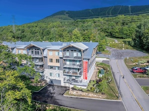 A multi-story residential building with balconies, a parking area, and a view of a forested hillside. - Mountain | Solstice Msa (Beaupré)