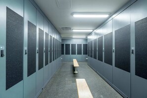A row of lockers with a bench in the center, a fluorescent light fixture on the ceiling, and a door with a number plate. - Mountain | Solstice Msa (Beaupré)