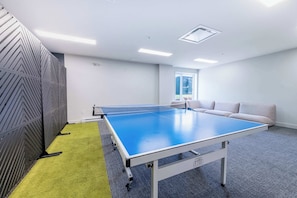 A ping pong table in a room with a grey carpet, a grey sofa, and a window. - Mountain | Solstice Msa (Beaupré)