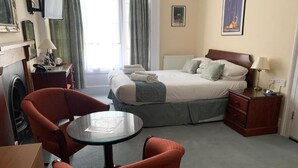 A hotel room with a bed, a round table, two chairs, a desk, a lamp, a television, and a radiator. - Peregrine House  (Canterbury)