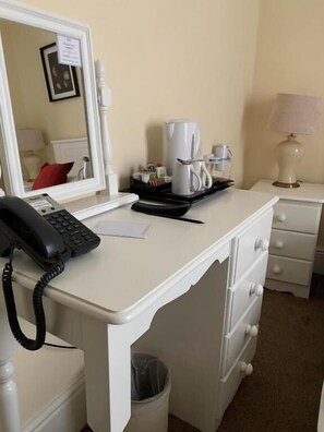 A hotel room with a white desk, a telephone, a coffee maker, a lamp, and a trash bin.