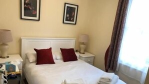 A hotel room with a bed, two bedside tables, a nightstand, a lamp, and a window with curtains. - Peregrine House  (Canterbury)