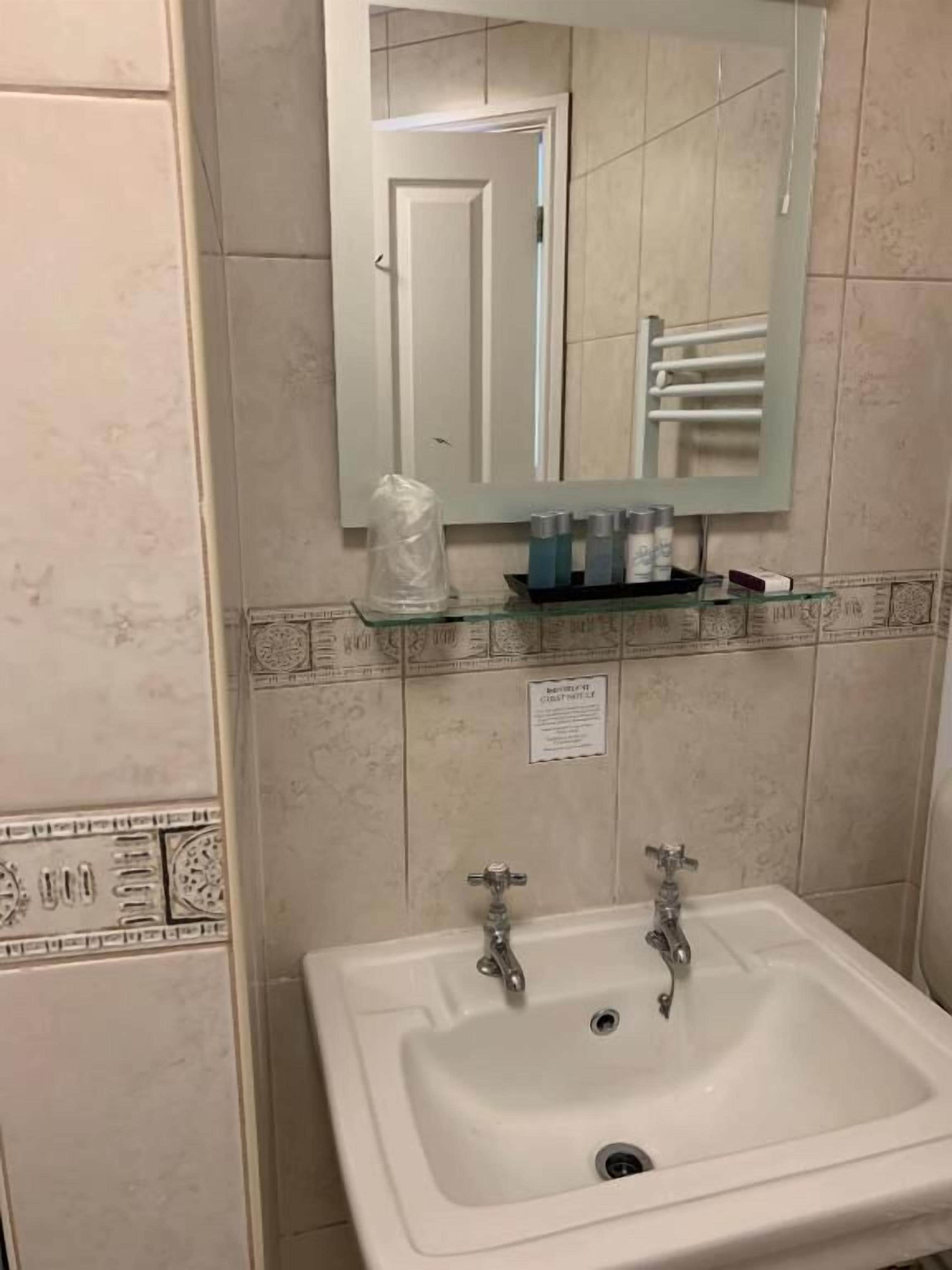 Une salle de bain avec un lavabo blanc, deux robinets, une étagère en verre où sont rangés les articles de toilette, et un miroir au-dessus du lavabo.