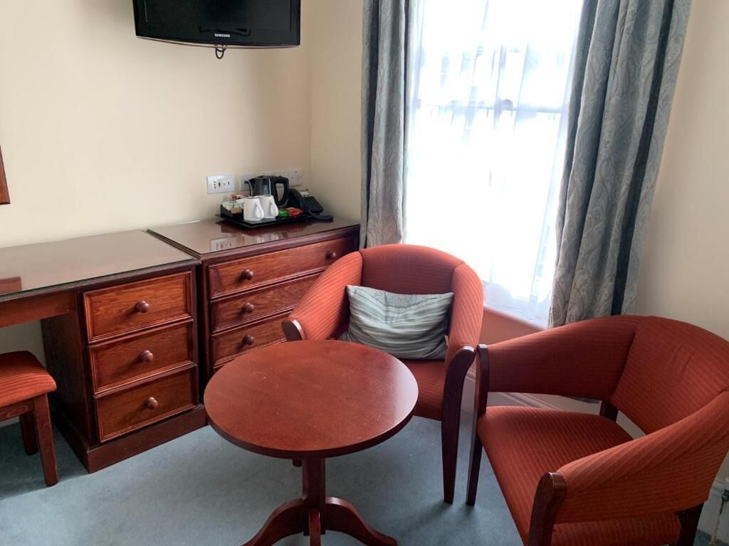A hotel room with a wooden desk, a chair, a small table, a television mounted on the wall, and a window with curtains.