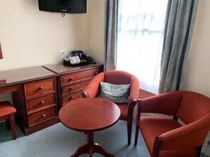 A hotel room with a wooden desk, a chair, a small table, a television mounted on the wall, and a window with curtains.