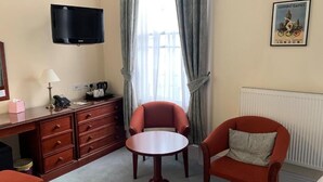 A room with a television mounted on the wall, a desk with drawers, a round table, two armchairs, and a radiator. - Peregrine House  (Canterbury)