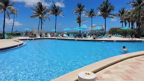 A swimming pool with lounge chairs and umbrellas, surrounded by palm trees. - The Tides Building (Hollywood)