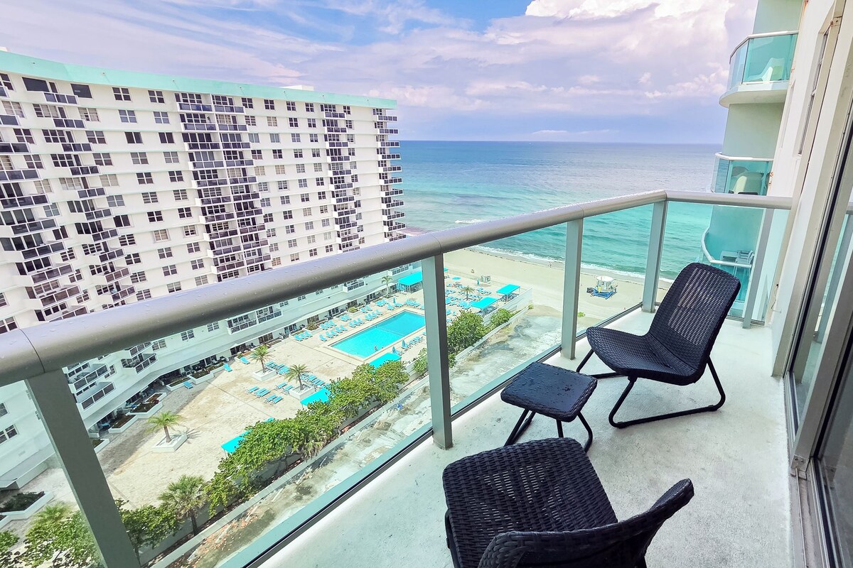 A balcony with two wicker chairs and a view of a beach with a pool.