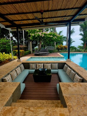 A covered outdoor seating area with a pool, surrounded by lush greenery and a bamboo roof.