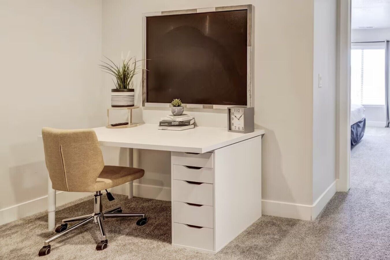 Un bureau blanc avec un écran d’ordinateur, une chaise et une horloge. On aperçoit une plante sur le bureau et une fenêtre à l’arrière-plan.